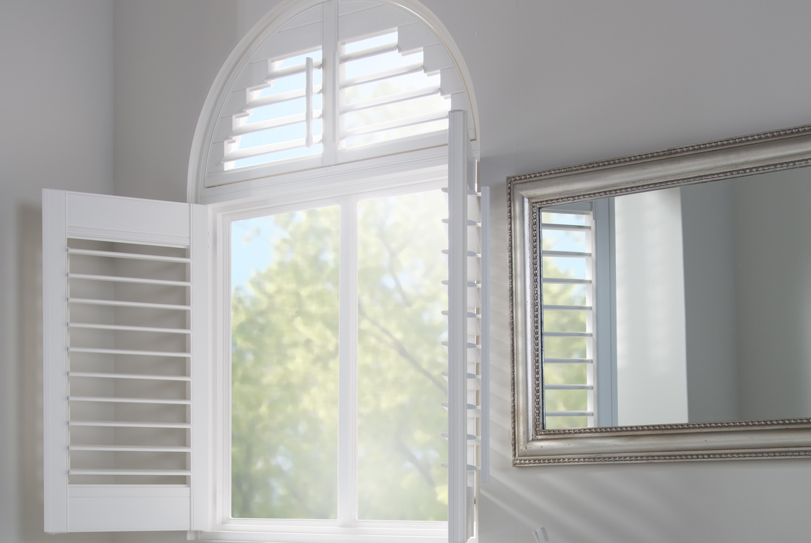 Off-white bathroom with arched window dressed in white shutters, lower shutters open, arched shutter at top closed with louvres open, mirror to right.