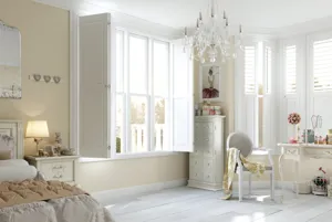 Classic French-style bedroom with cream walls, white painted wood floor, bed, dressing table, and chest of drawers in white classic style, chandelier lighting and two sets of windows dressed in white shutters with solid lowers and louvred uppers, partially open.