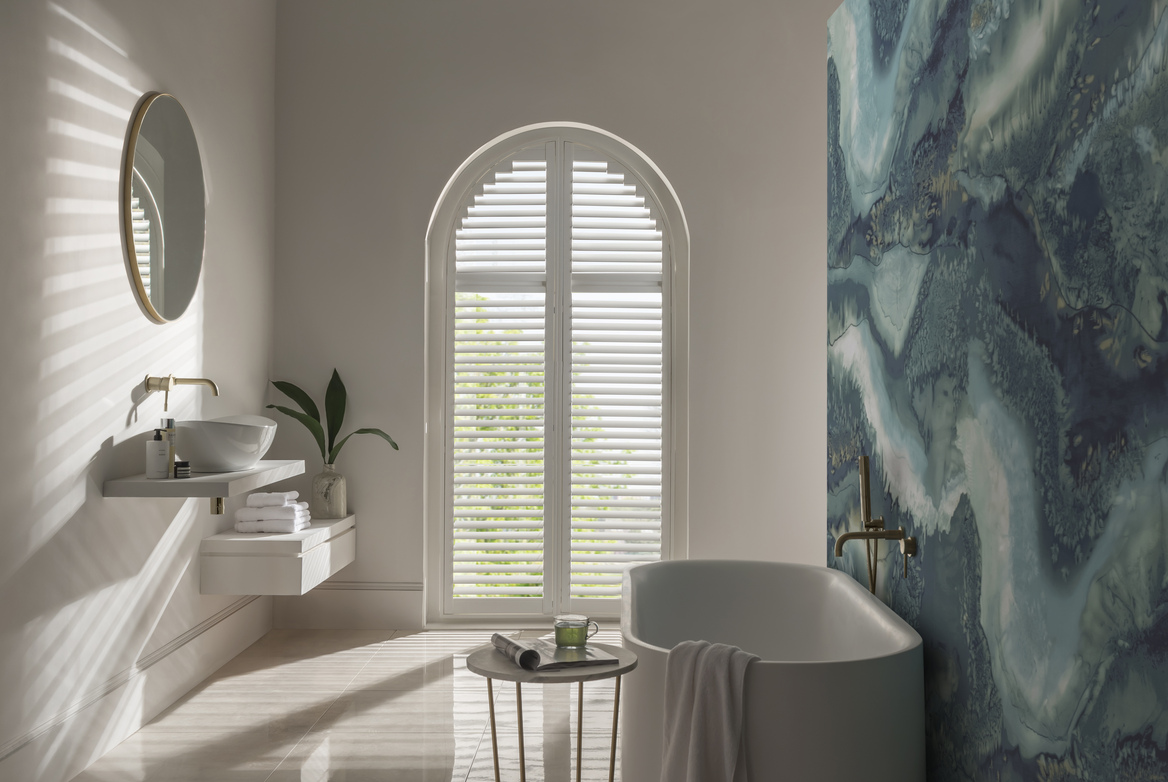 White bathroom with bath on right against feature wall with abstract marble-effect in blues and arched window to rear dressed in white shutters, louvres open.
