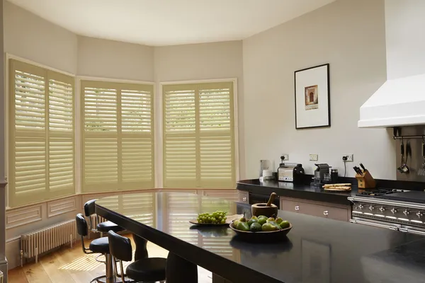 Cream kitchen with bay windows dressed in light ochre full height shutters, closed with louvres open, kitchen units with black worktops, island dining  unit with black worktop and black stools.