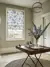 Hallway with wooden floor with round jute rug under folding card table with vase of blue flowers, white stairs to left and window dressed in roller blind with large blue flowers and green foliage