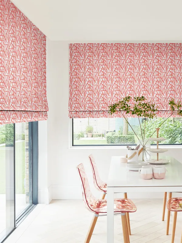 Dining room in white with window to rear and patio doors to left dressed in Roman blinds with orange-red geometric linear design on white background, with modern white table and pink marl chairs with pink and white crockery and vase with tall stemmed greenery.