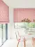 Dining room in white with window to rear and patio doors to left dressed in Roman blinds with orange-red geometric linear design on white background, with modern white table and pink marl chairs with pink and white crockery and vase with tall stemmed greenery.