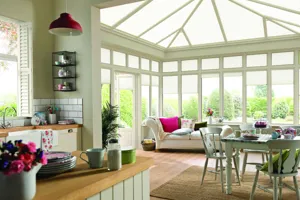 Country chic open plan kitchen/diner to conservatory with wood floor, brown rug under pale olive painted wood table and chairs, white chaise lounge in background with side and roof windows dressed in alabaster duette blinds at varying heights.