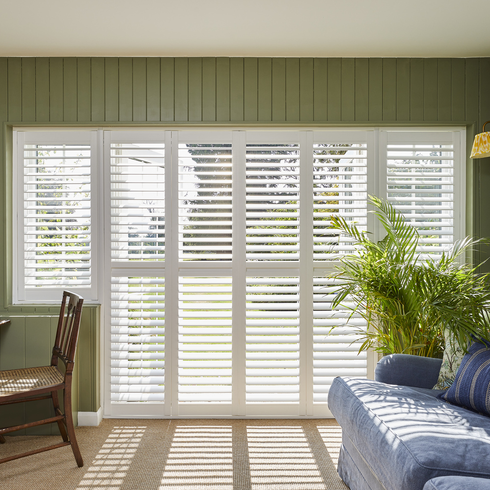 Room with windows and doors dressed in white tracked door shutters partially open. Sofa to right, desk and chair to left.