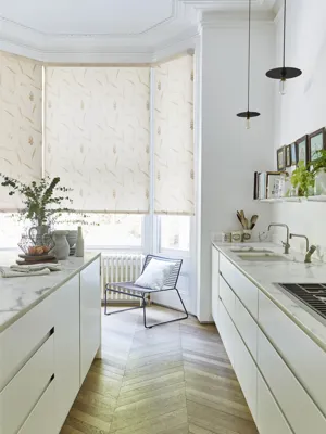 Kitchen in white with cream units and pale parquet floor, marble worktops and bat window dressed in roller blind in cream with wheatsheaf pattern.