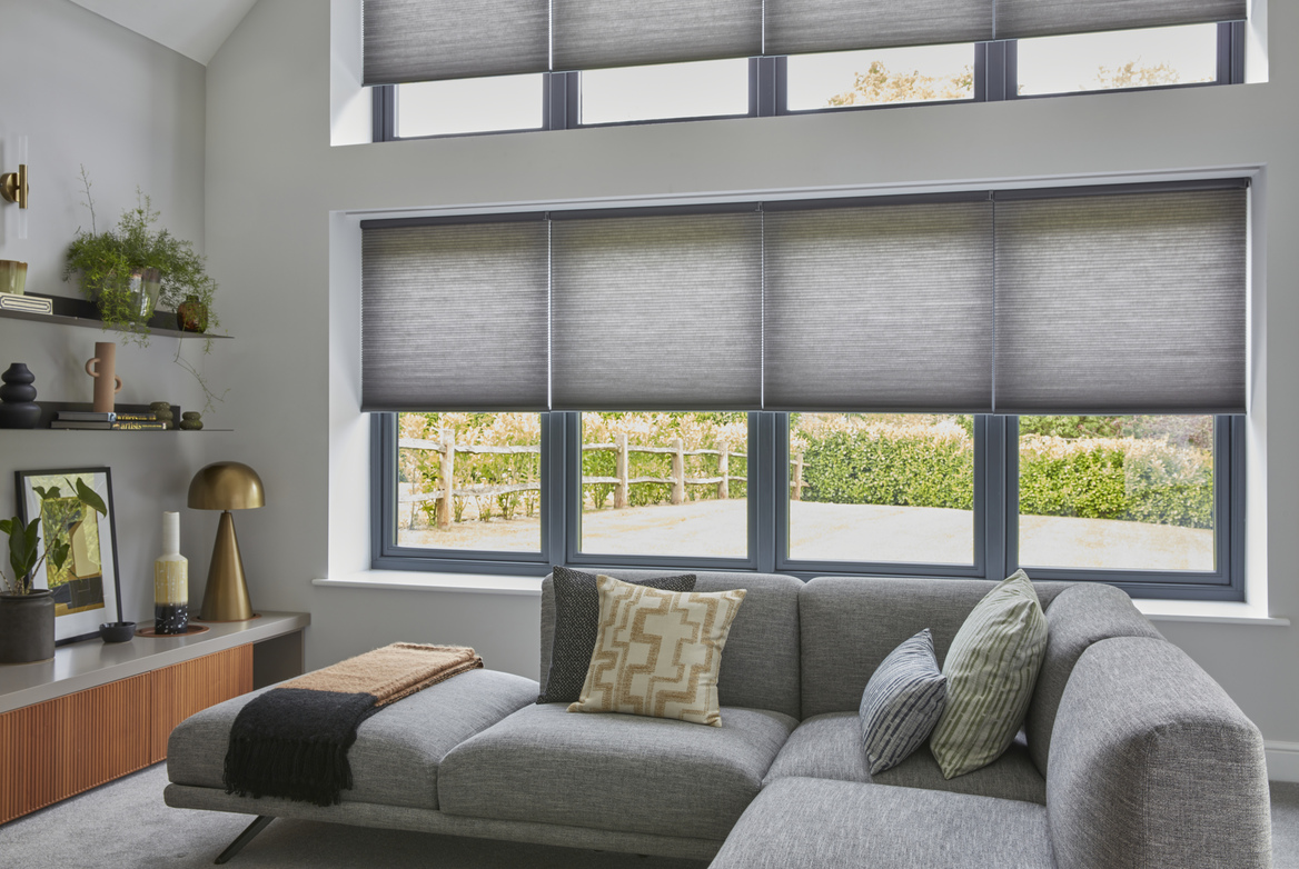 Pale grey living room with to sets of four windows one above the other dressed in pale grey duette blinds lowered halfway, L-shaped grey sofa with cushions in chartreuse and blue patterns.