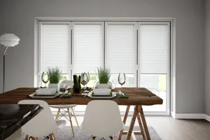 Dining room in pale grey with pale wood floor, wooden trestle-style table with white contemporary chairs, table set with white crockery and bifold doors dressed with white blinds within frame, doors closed