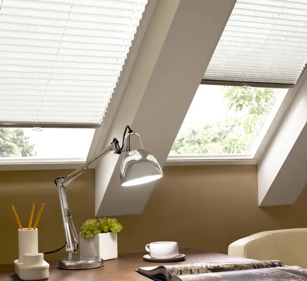 Two adjacent skylights in an eave recess dressed in sheer white pleated blinds with a desk underneath with sundry stationery accessories.
