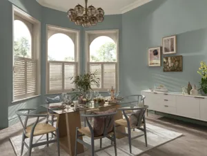 Dining room with three windows on a curved wall dressed in white cafe style shutters, closed with louvres open, pale blue walls, modern glass dining table with wood and metal chairs, white low sideboard.