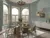 Dining room with three windows on a curved wall dressed in white cafe style shutters, closed with louvres open, pale blue walls, modern glass dining table with wood and metal chairs, white low sideboard.