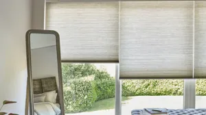 Close up of bedroom with standing mirror in front of windows and doors dressed in beige-grey duette blinds at varying heights.