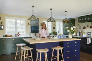 White shutters fitted to rectangular windows in a kitchen with yellow walls, green and blue cabinets and breakfast stools