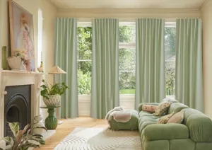 Cream living room with wall of windows dressed in four pale green curtains, open, matching sofa opposite a cast iron fireplace with cream surround, large modern portrait on mantlepiece, cream rug on a pale wooden floor.