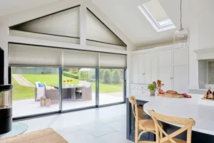 White gable end kitchen diner with white tiled floor, white units to the side, island dining area with blue units and white worktop and skylight, gable end windows and doors all dressed in mid grey duette blinds, skylight blind three quarters lowered, gable shape fully lowered and door blinds partially lowered.