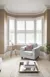 Neutral lounge with cream carpet and rug, pale grey chair in a curved 5-bay window dressed in white shutters, louvres open.