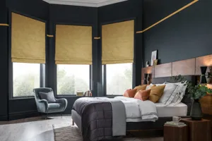 Large bedroom with wooden floor, bay window with three ochre faux velvet Roman blinds, navy walls, bed with white bedding and grey quilted throw,. 