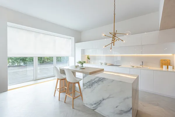 Open-plan white kitchen with trio of window and doors dressed in white roller blind with marble island with white chairs and modern pendant light.