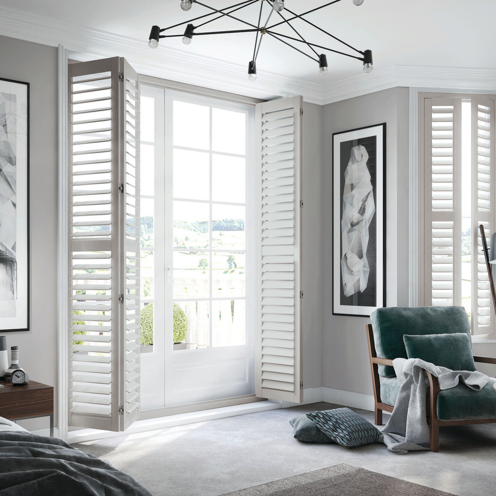 full height tracked white vinyl shutters, folded back in a modern style bedroom with large black and white pictures ,pale grey floor, and a dark grey lounger.