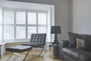 Living room with pale wood floor, black leather sofa, black table lamp on silver square end table, black leather chair and footstool in a box bay window dressed in white shutters, closed, louvres open.