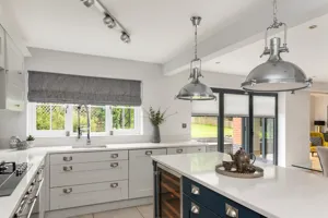 Kitchen part of open-plan space with pale grey units top and left, with white marbled workstop, blue island unit on right with same worktop and window dressed in grey Roman blind, partially lowered.