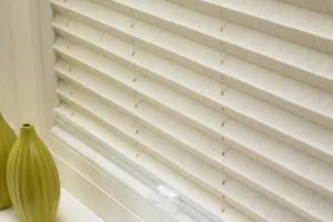 Close up of an ivory pleated blind with a feint threaded pattern in a slightly deeper shade, fully lowered in a window recess with a tab control, two small olive conical stem vases on the sill.