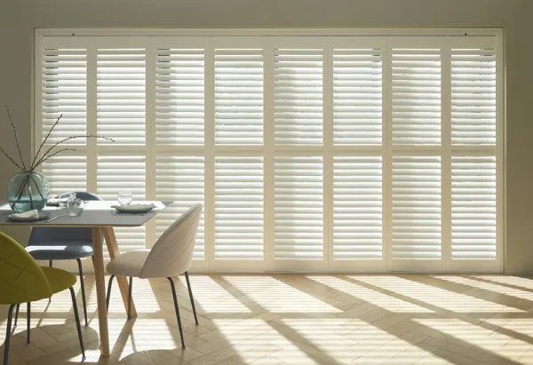 Dining room with contemporary table and chairs on left and large expanse of doors and windows dressed in white tracked shutters, closed with louvres open and light rays on the floor.