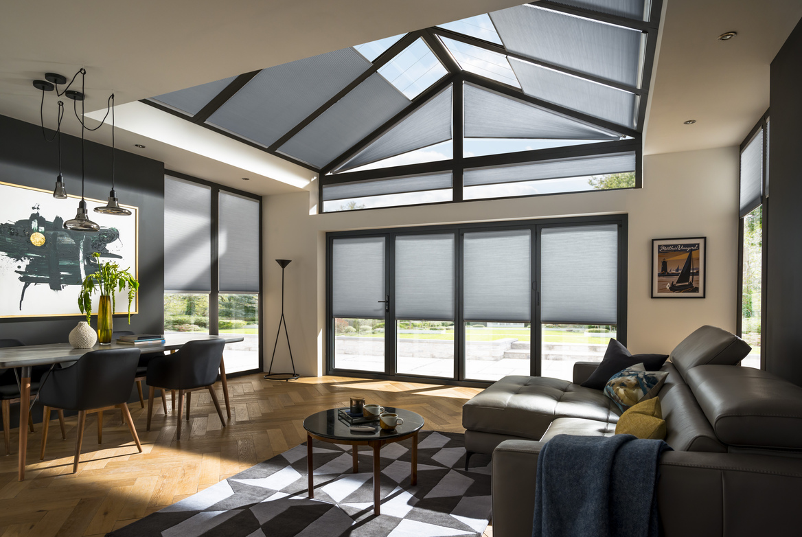 Open plan lounge diner with gable end and roof lantern windows and doors dressed in pale grey duette blinds, parquet floor, chocolate brown sofas on right, dining table and chairs on left with a geometric white and dark grey patterned rug in the middle.