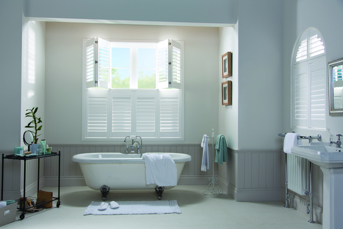 White bedroom with window in alcove dressed in tier on tier white shutters, top half partially open, bottom half closed, louvres open on both above white bath, arched window on right dressed in white shutters closed with louvres closed.