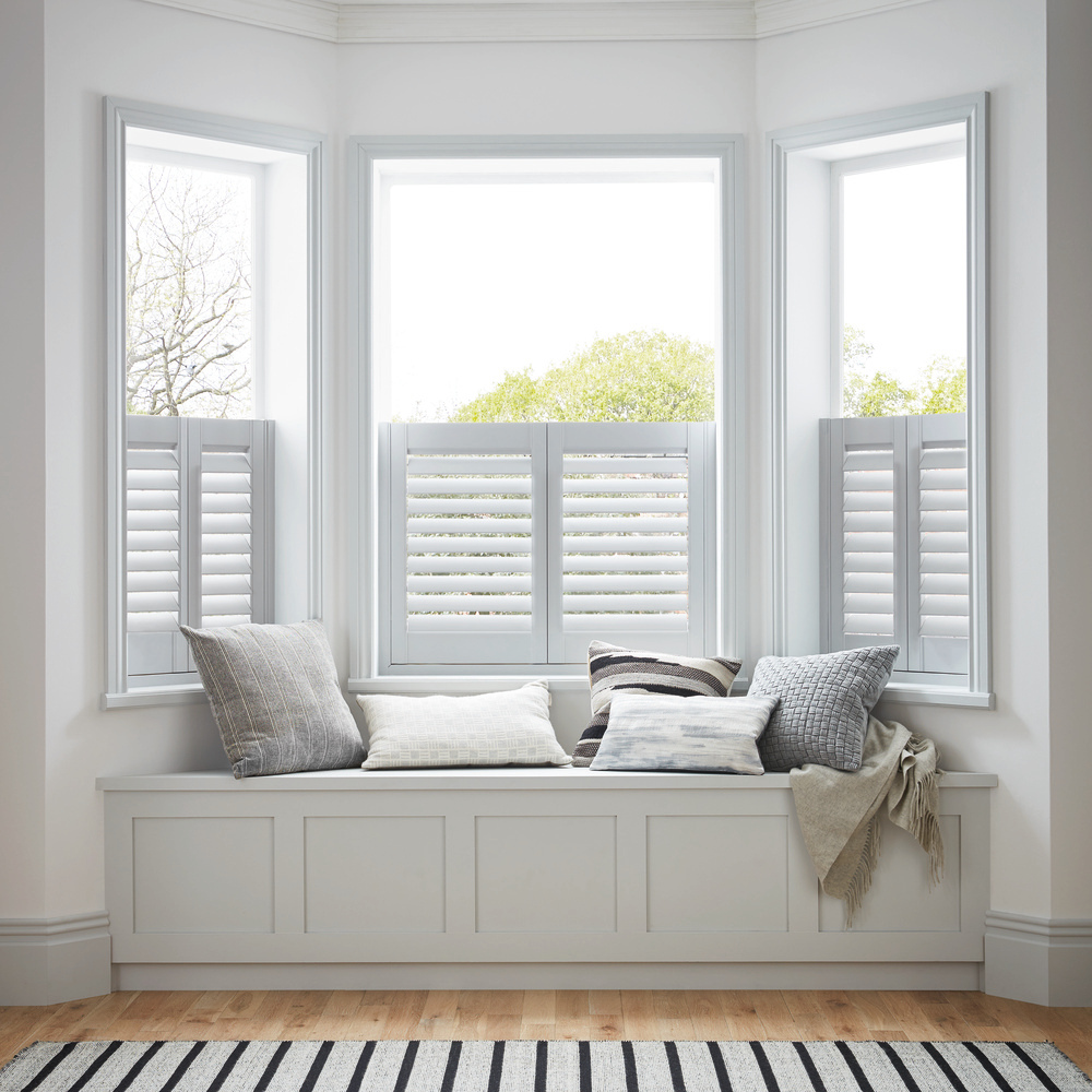 White cafe style shutters, closed with louvres open in a white room with pale wood floor, white rug with blue stripes in front of a bay window with a window seat filled with white and pale grey cushions.