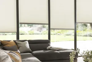 Close in on a brown L-shaped sofa in front of floor to ceiling windows and doors dressed in pale ivory duette blinds at varying heights.