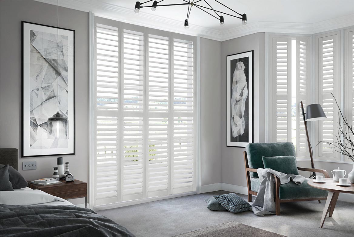 In a bedroom that has a bed, sidetable, armchair and coffee table are Arctic Horizon Shutters. The White coloured Shutters are fitted to a series of tall windows in the bedroom which is decorated in neutral grey tones.