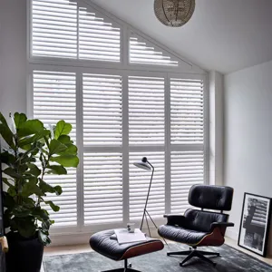 White lounge with black recliner chair and footstall in front of windows and doors with additional angled window to the top all dressed in white shutters, louvres open.