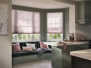 Contemporary kitchen with banquette seating under bay windows dressed in soft pink Duette® blinds drawn halfway, co-ordinating cushions on blue seating, kitchen counters in soft grey with white top.