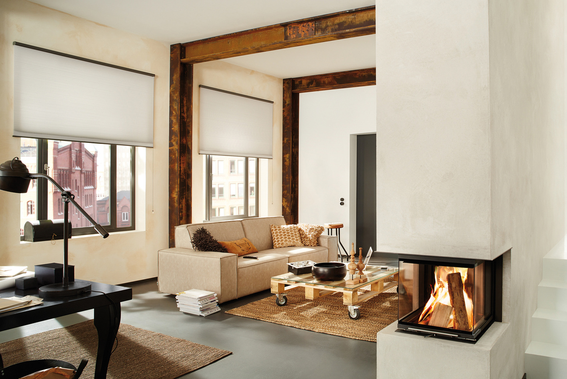 Loft apartment style lounge room with beams and feature pillar fire, lit. and two large windows dressed in pale Duette blinds slightly lowered,