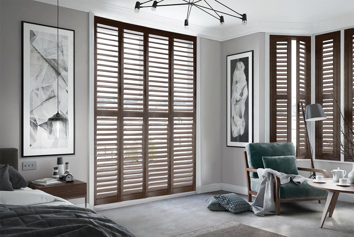 Burley Burnished Coal Shutters have been fitted to tall windows in a bedroom. The Shutters are Natural, Black coloured while the bedroom is decorated with grey tones.