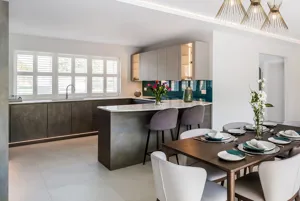 White kitchen diner with L-shaped dark wood units, table, white chairs, and windows  dressed in wite shutters, closed with louvres open.