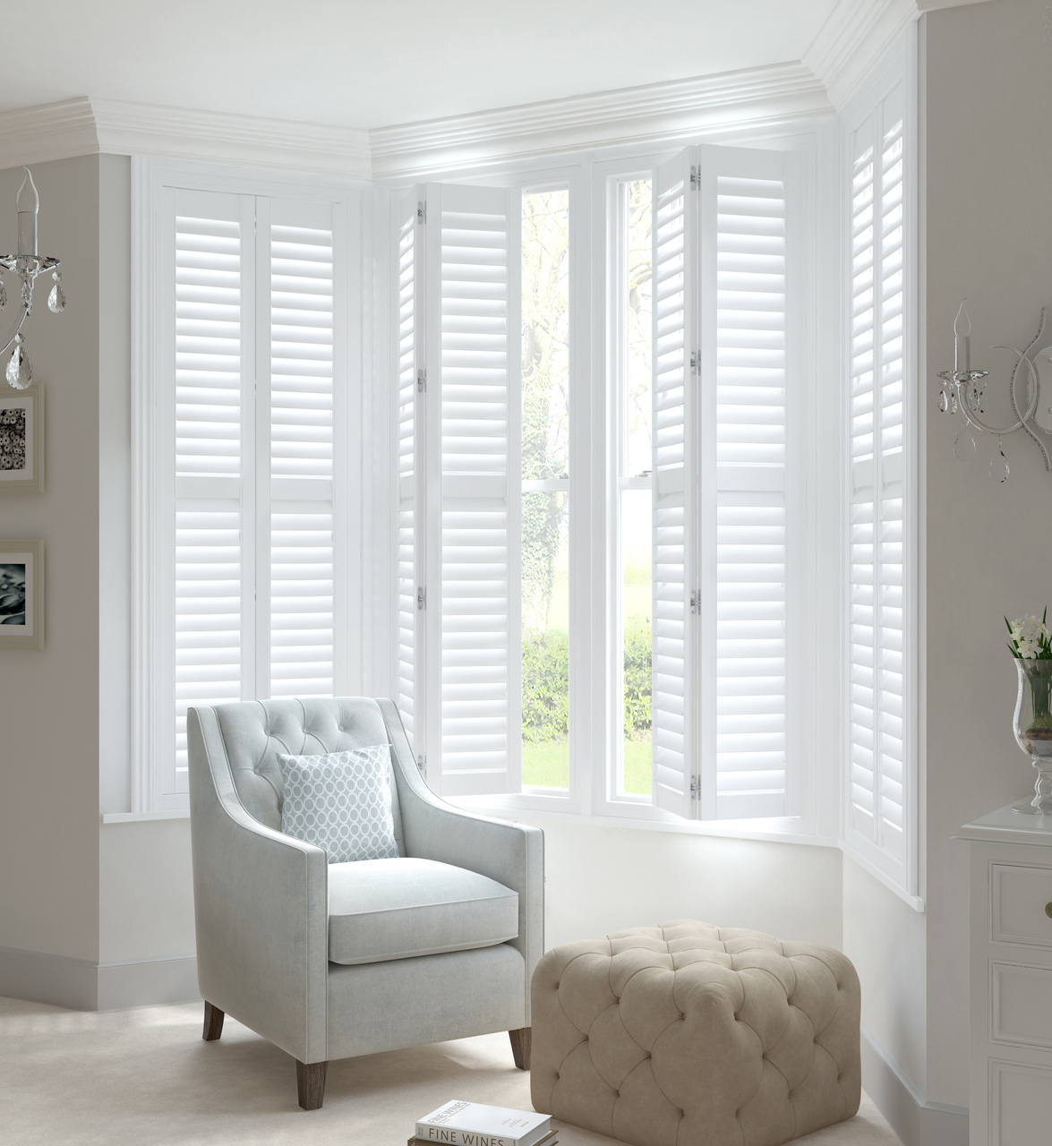 Living room in pale grey with white full height shutters in a bay window, partially open, louvres open, pale grey chair in recess, pale beige pouffee in front of chair.