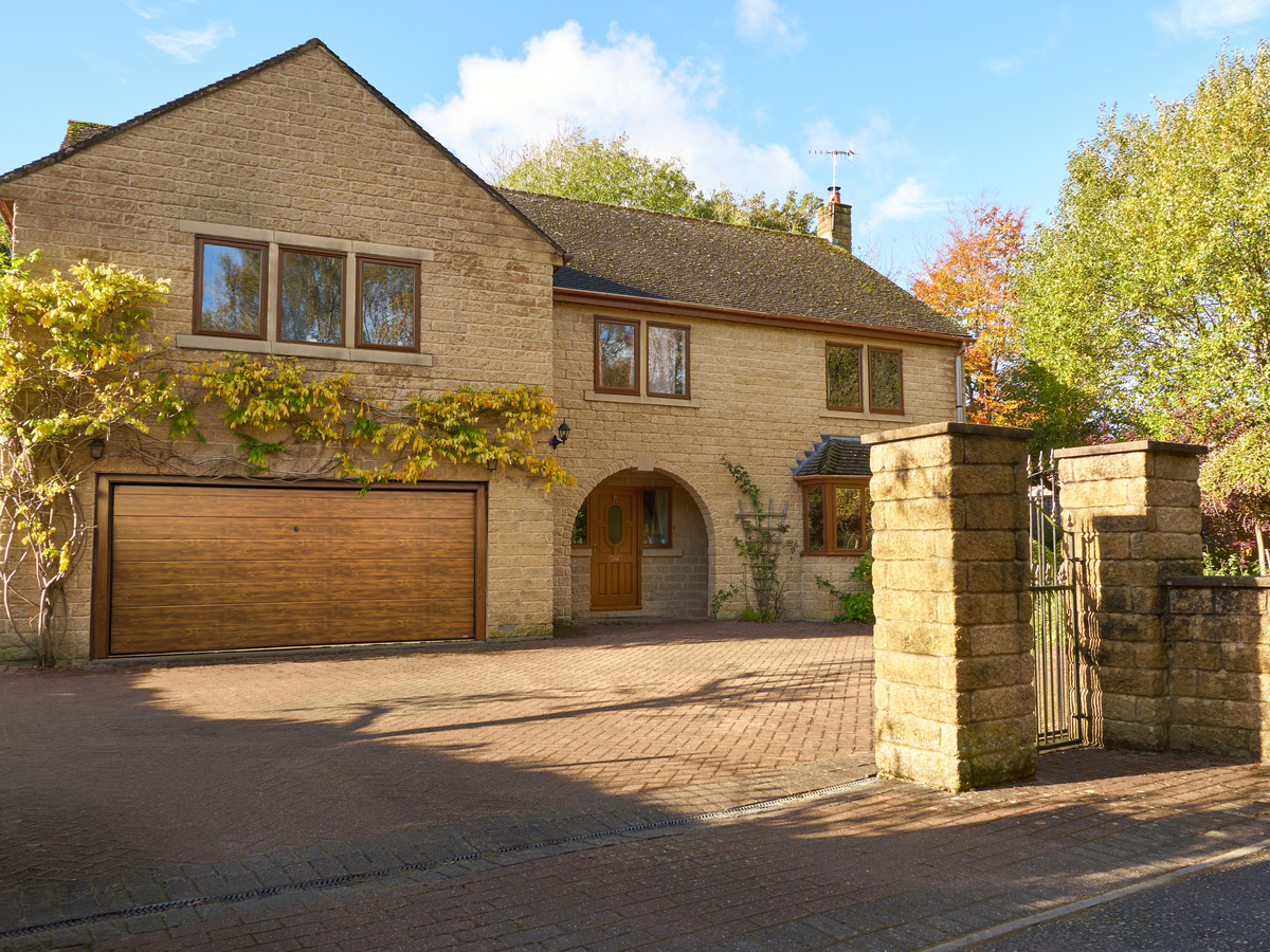 Large Yorkshire-style stone house and drive with integral garage door in mid brown matching front door, with climbing plant above garage.