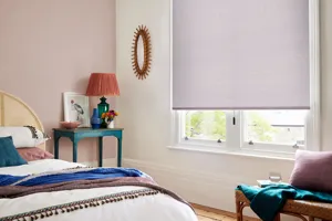 Bedroom with window dressed in plain lilac roller blind., soft pink and white walls, wood floor with cobalt blue rug with russet fringe, white bedding with bead detail.