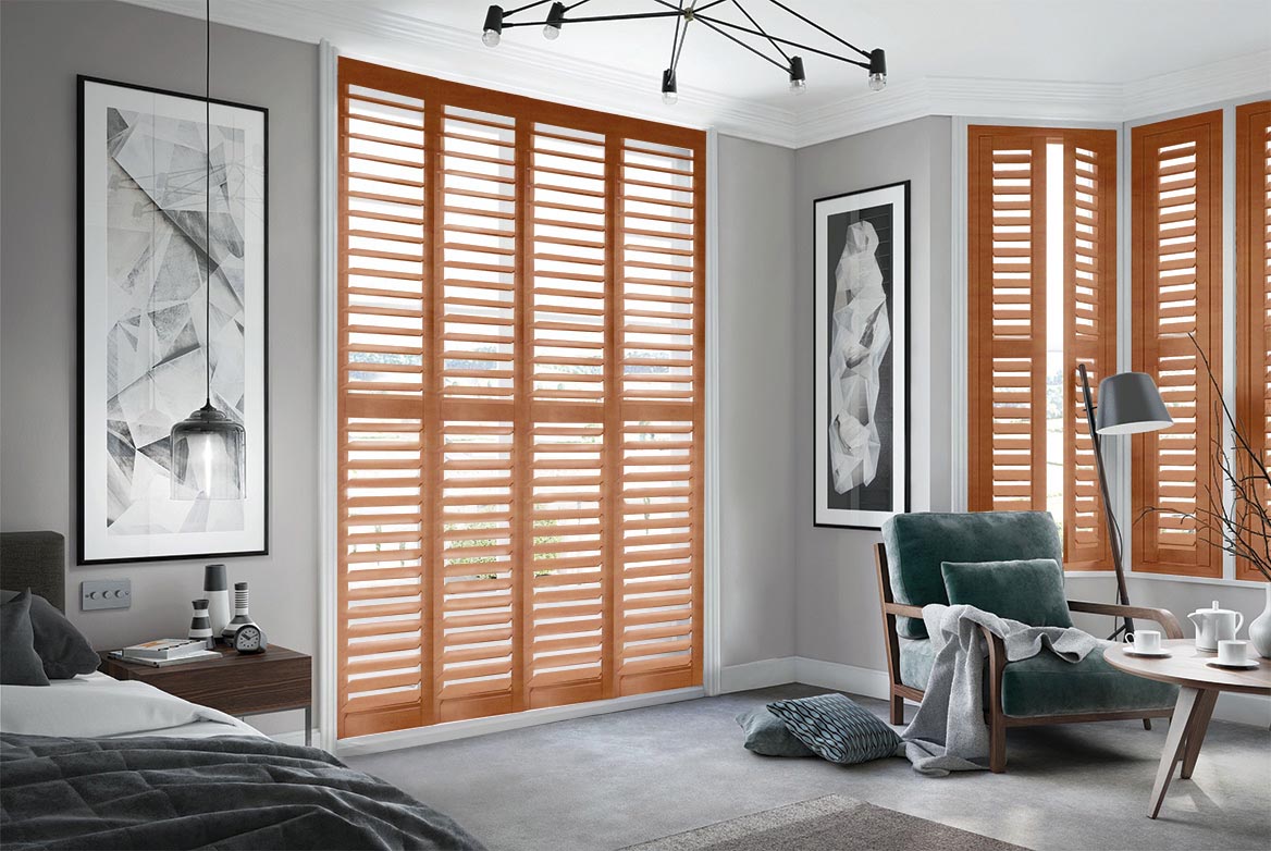In a bedroom that is decorated in tones of grey Burley Tennessee Oak Shutter have been fitted to tall windows. The bedroom features a bed, side table, armchair and coffee table