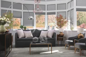 Extended conservatory with grey flooring, grey rug, dark grey upholstered sofa and chairs, low table with glassware, pale pink cushions, low-hanging pendant light with wirework shades and sides and roof dressed in pale grey duette blinds.