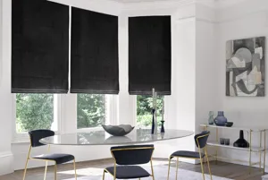 Modern dining room with oval glass table, metallic gold chairs with blue upholstery, metallic gold side table with blue glassware and bay window dressed in three navy blue velvet Roman blinds.