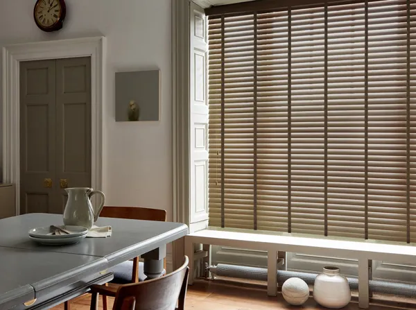 Dining room in soft white with large window dressed in mid brown wooden venetian blinds, lowered, slats tilted, grey table  and door, white bench in window recess with white ceramic vase underneath.