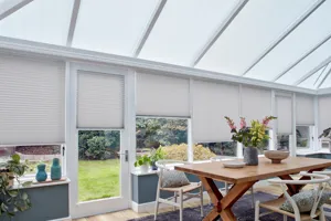 Rectangular conservatory with wooden dining table, white chairs and pale grey pleated blinds on side windows and off-white blinds in roof.
