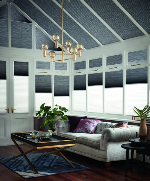 Conservatory with side windows in twinshade duette blinds dark grey on top and white beneath, all at varying heights, dark grey roof blinds., dark wood floor, pale grey sofa.