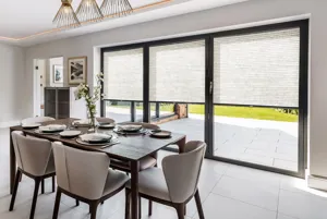 Dining room with pale mink walls, large tiled floor, wooden dining table and upholstered chairs in pale mink, with three bi-fold doors dressed in natural duette blinds, lowered halfway.
