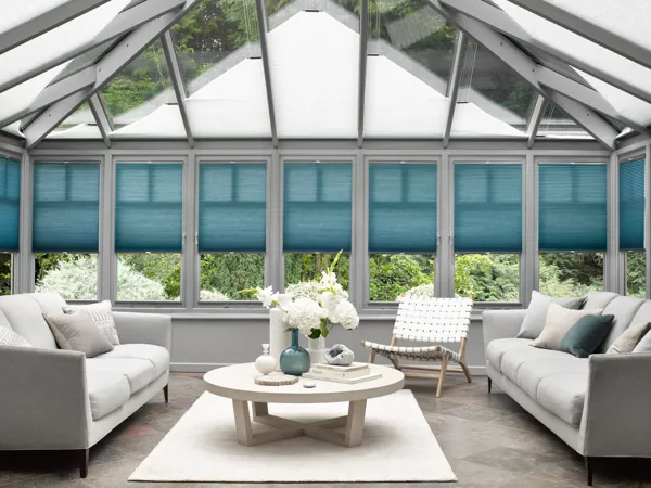 Large conservatory in pale grey with side windows dressed in marine blue duette blinds lowered halfway, roof windows dressed in grey duette blinds at varying heights, mid grey floor, white rug under a blonde table with white accessories and flowers, white-grey sofas facing each other.