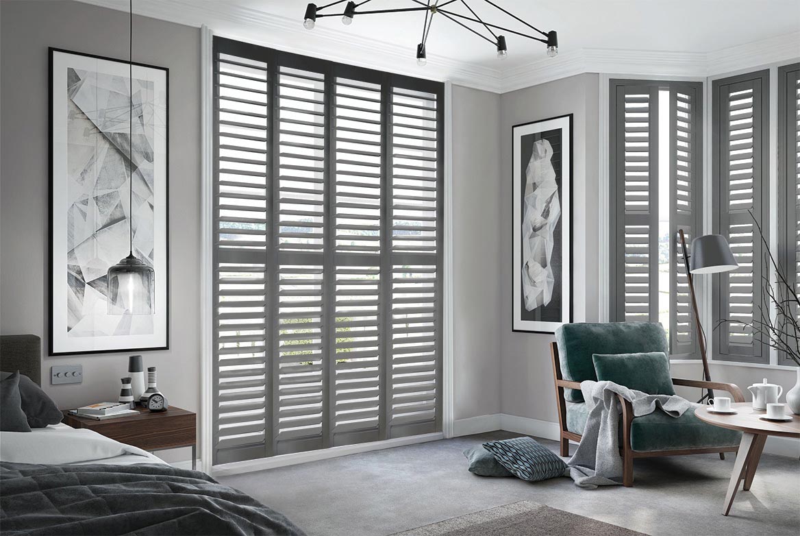 In a bedroom that has a bed, sidetable, armchair and coffee table are Midnight Forge Shutters. The Grey coloured Shutters are fitted to a series of tall windows in the bedroom which is decorated in neutral grey tones.