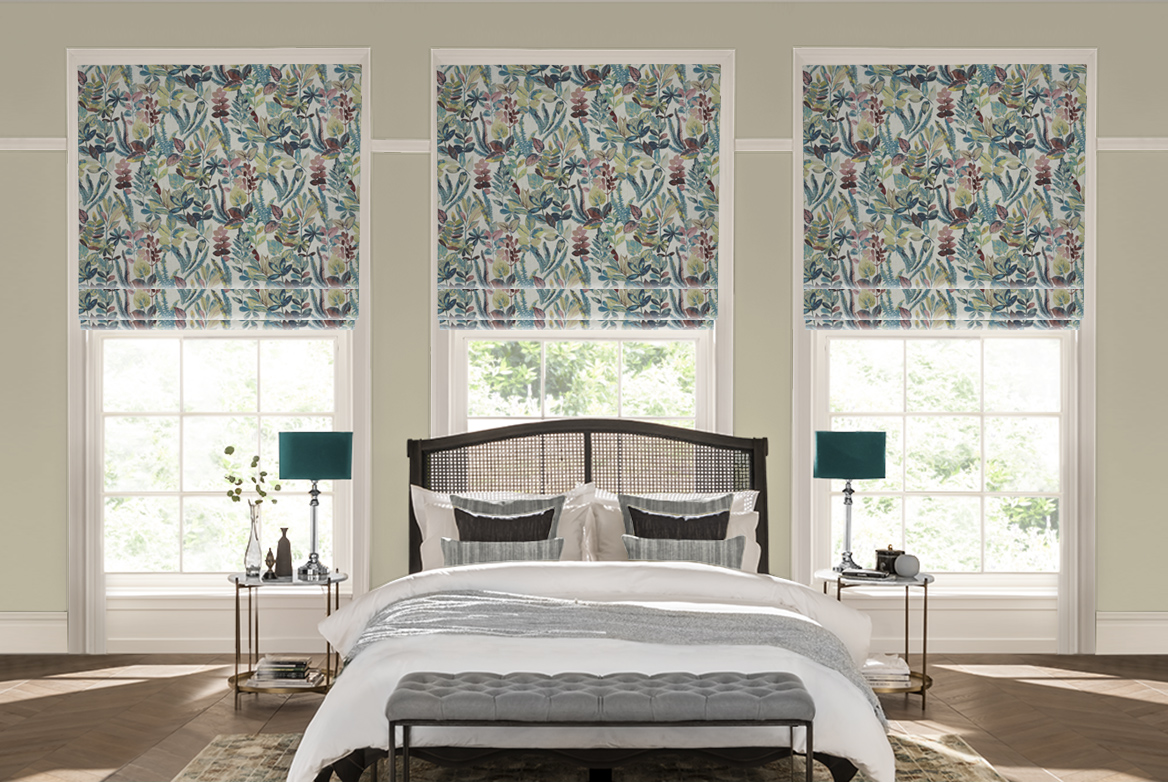 Bedroom with wood floor and large brown rug under double bed with mesh headboard, two bedside tables with candlestick lamps with teal shades, white bedding with silver throw and silver and black cushions, grey footstool at bed end and three windows dressed in white with abstract flower and leaf patterned Roman blinds.